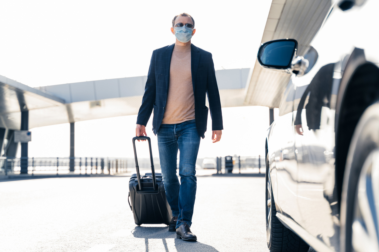 Passenger wearing mask pulling suitcase towards waiting chauffeur car at airport